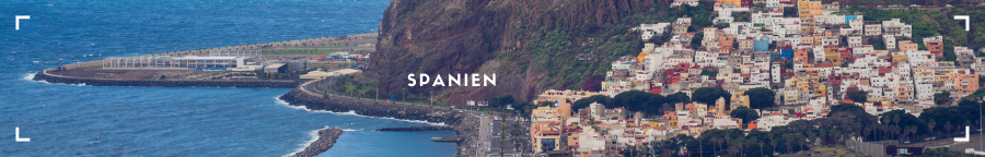 Eine malerische Stadt an der spanischen Küste, die sich direkt am Meer erstreckt. Im Vordergrund sind bunte Häuser auf Hügeln zu sehen, die sich zum Wasser hinunterziehen. Die Uferpromenade ist belebt mit Menschen, die die Sonne genießen, und kleine Boote schaukeln sanft auf den Wellen.
